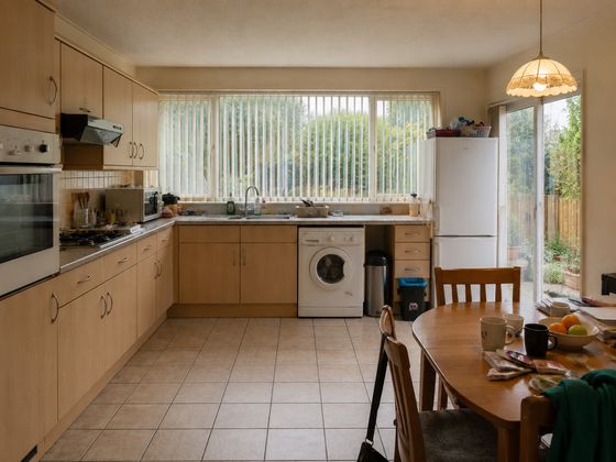 Dated 1990s update of a 1970s British semi-detached kitchen-diner with beech-effect laminate units, freestanding appliances, patio door and cluttered dining table before makeover.