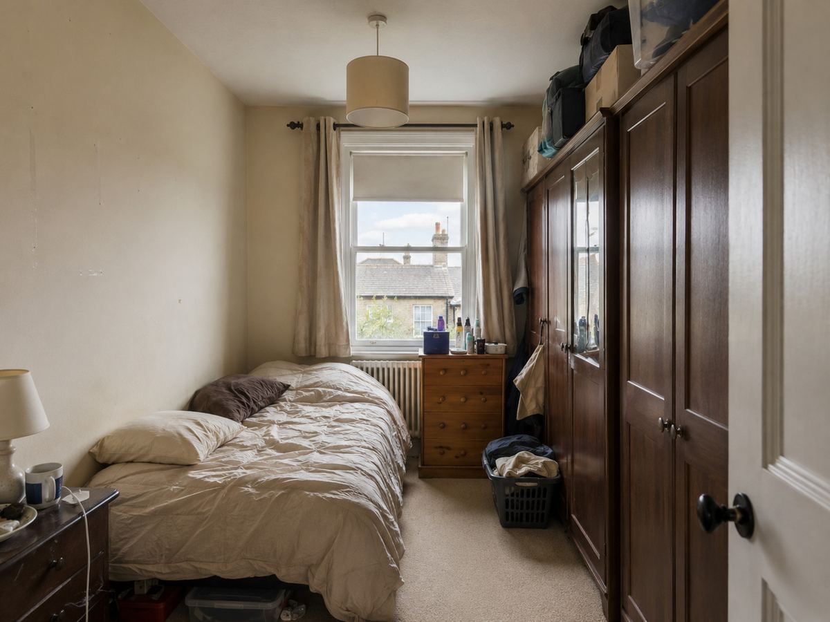 Dated compact British bedroom with a divan bed, old wardrobe, plain walls, radiator and uPVC window before redesign.