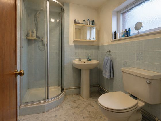 Small dated British shower room with a quadrant shower, pedestal basin, toilet, pale blue-grey tiles and old vinyl flooring before its makeover.